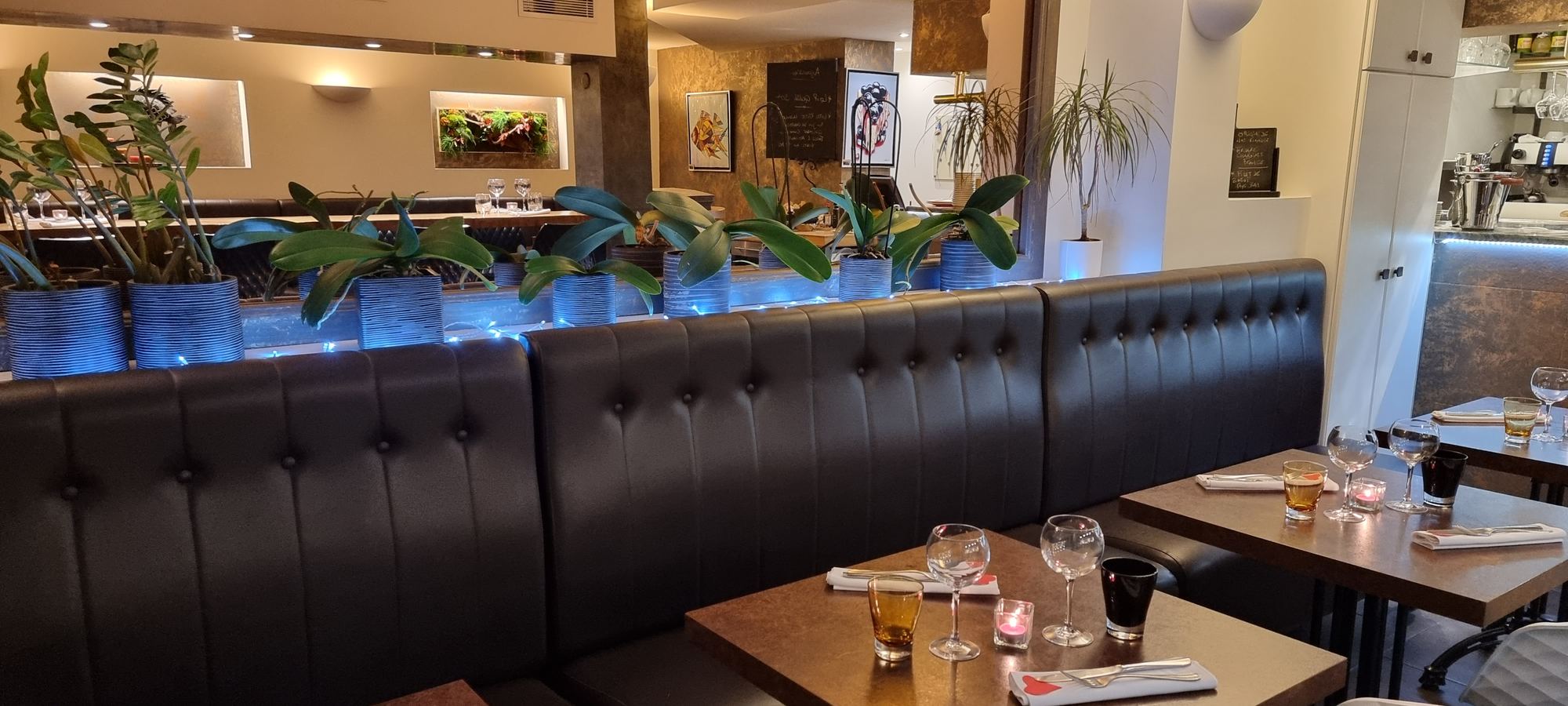 La salle : banquette capitonnée en cuir noir, tables en bois, plantes et décor marin